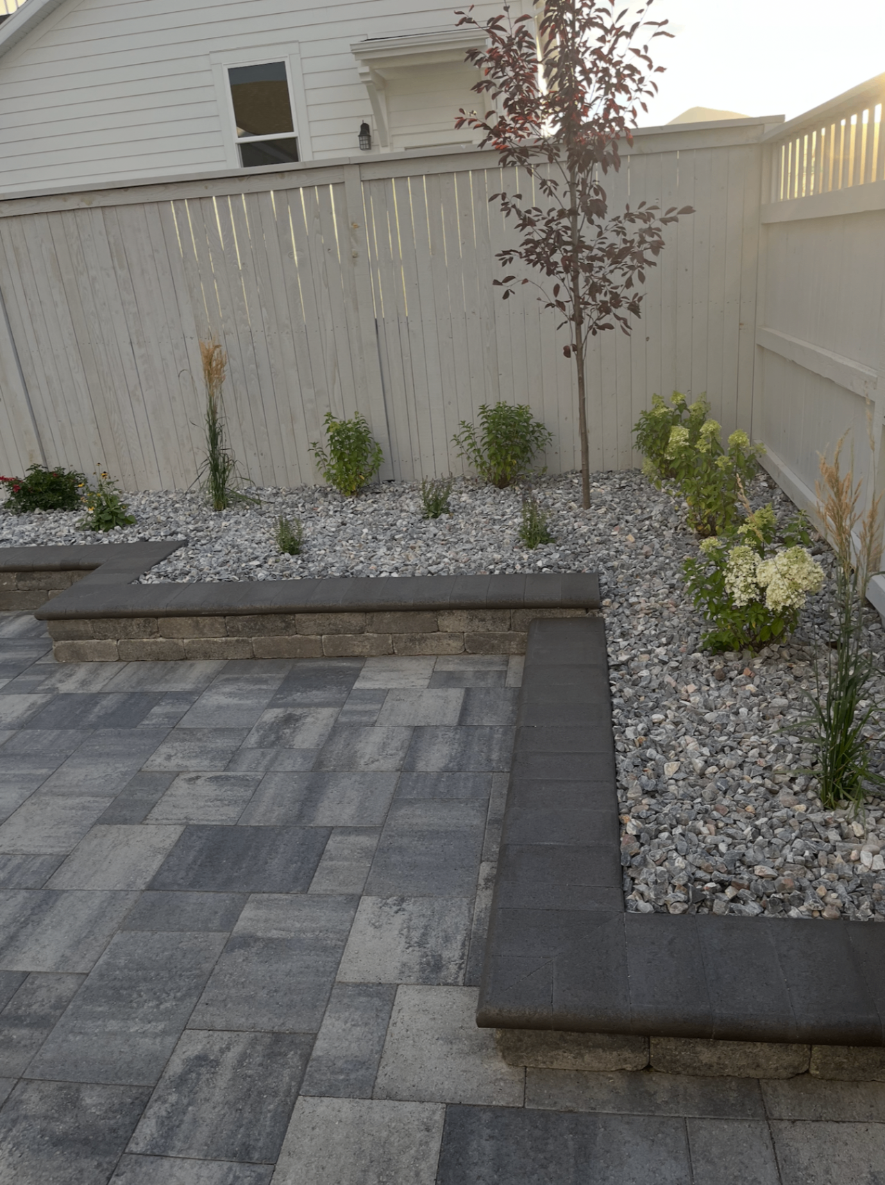 Paver patio with raised planter bed, decorative rock, and young trees